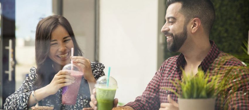 Dois amigos, um homem e uma mulher, com copos de sucos coloridos na mão e rindo
