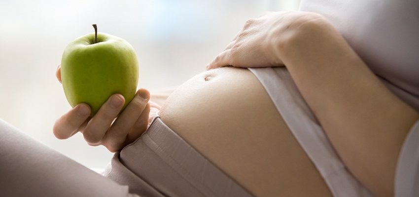 Foto de uma mulher grávida deitada, com a mão na barriga, segurando uma maçã verde.