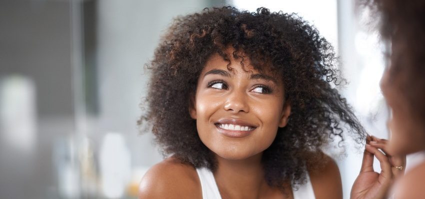 Foto de uma mulher feliz, de camiseta branca, olhando no espelho o seu cabelo cacheado.