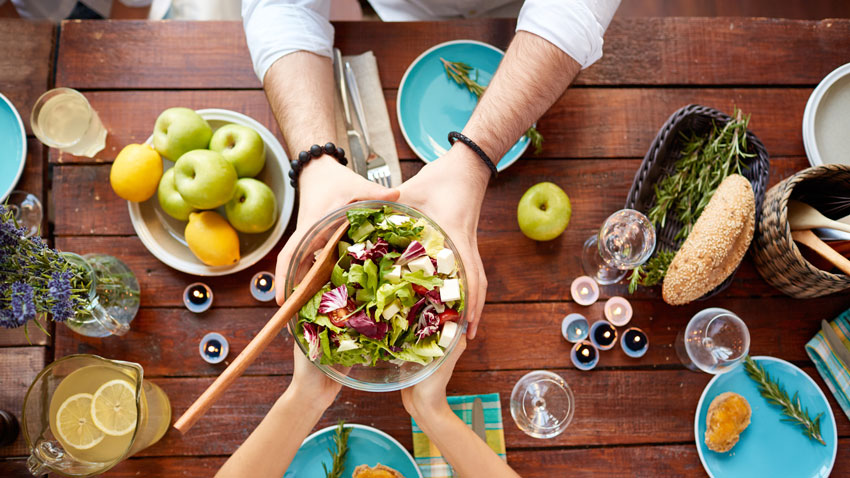 Salada sendo segurada por duas pessoa em uma mesa com alimentos saudáveis.