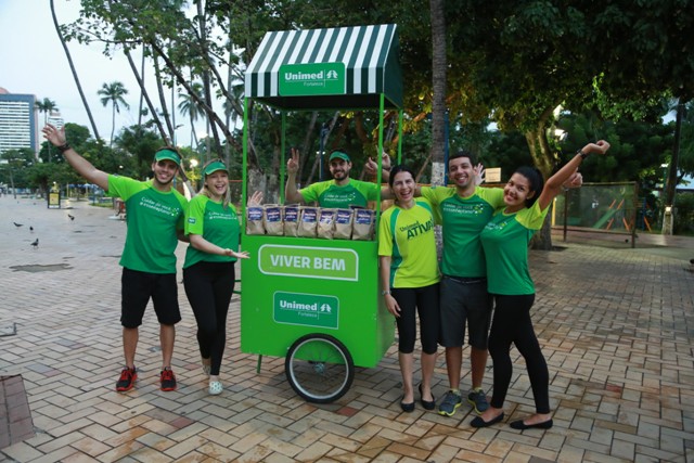 Colaboradores na Beira-Mar de Fortaleza, com os braços levantados, ao lado de um carrinho verde, onde se lê "viver bem", que estavam utilizando para distribuir frutas aos passantes.