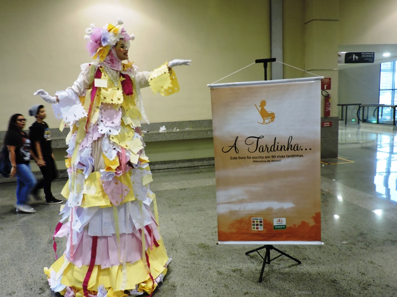 Mulher com vestido bem colorido ao lado do cartaz do livro "A Tardinha"