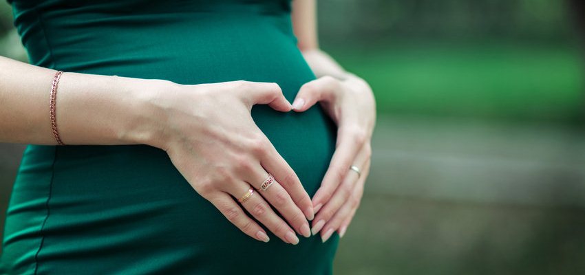 Mulher grávida vestida de verde fazendo um coração com as mãos na barriga.