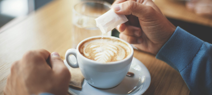 Mão colocando açúcar em uma xícara de café para causar reflexão em como reduzir o consumo