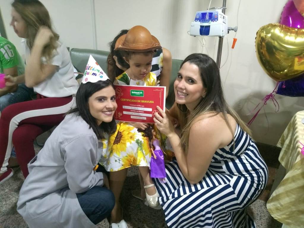 Uma enfermeira e a me de Giovanna segurando o certificado de fim do tratamento com Giovanna ao fundo.