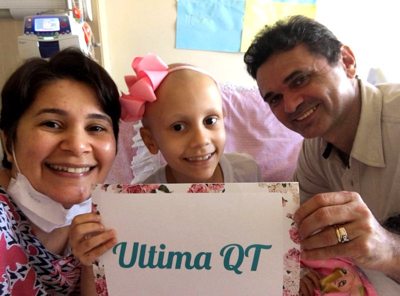 Maria Alice sorridente segurando um cartaz com o texto "Ultima QT" ao lado de seus pais